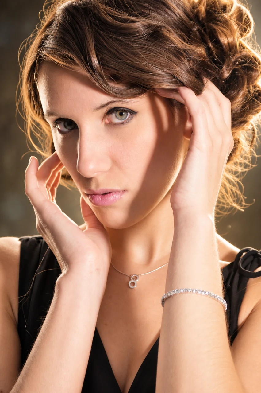 Close-up portrait of a brown-haired woman with styled curls, both hands framing her face, wearing a black top, silver necklace, and bracelet under warm studio lighting.