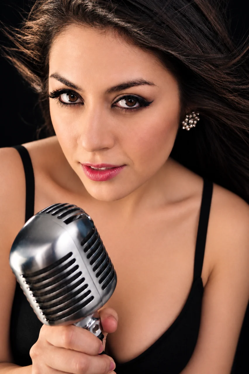 Close-up portrait of a dark-haired woman holding a large vintage silver microphone, wearing black with jeweled earrings and dramatic eye makeup against a dark background.