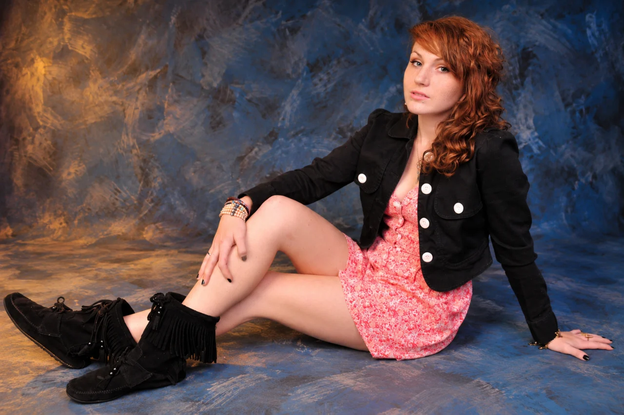 A red-haired woman reclines on a blue studio floor, wearing a pink patterned dress, cropped black jacket, and black fringed boots. One knee is raised while her other hand supports her against the floor.