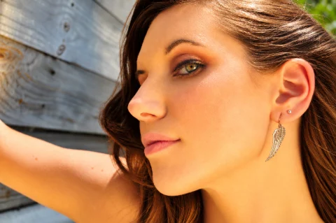 Close-up side-profile portrait of a brunette woman with hazel-green eyes, soft pink lips, and a dangling silver angel-wing earring. Warm sunlight illuminates her face and hair as she poses with one arm raised against a weathered wooden wall.