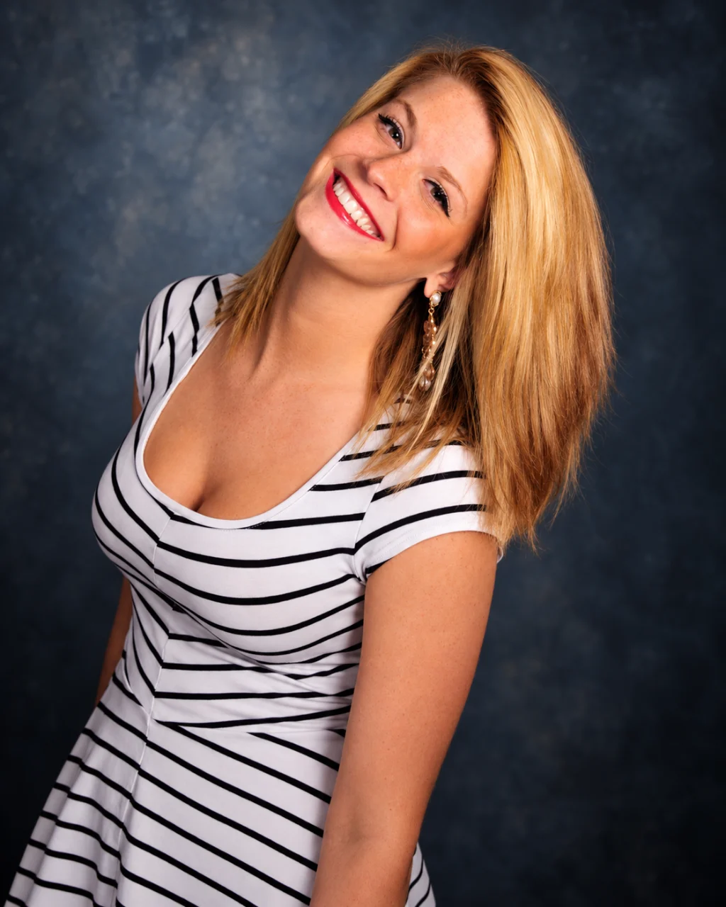 A smiling blonde woman leans toward the camera in a black-and-white striped dress against a blue studio backdrop.