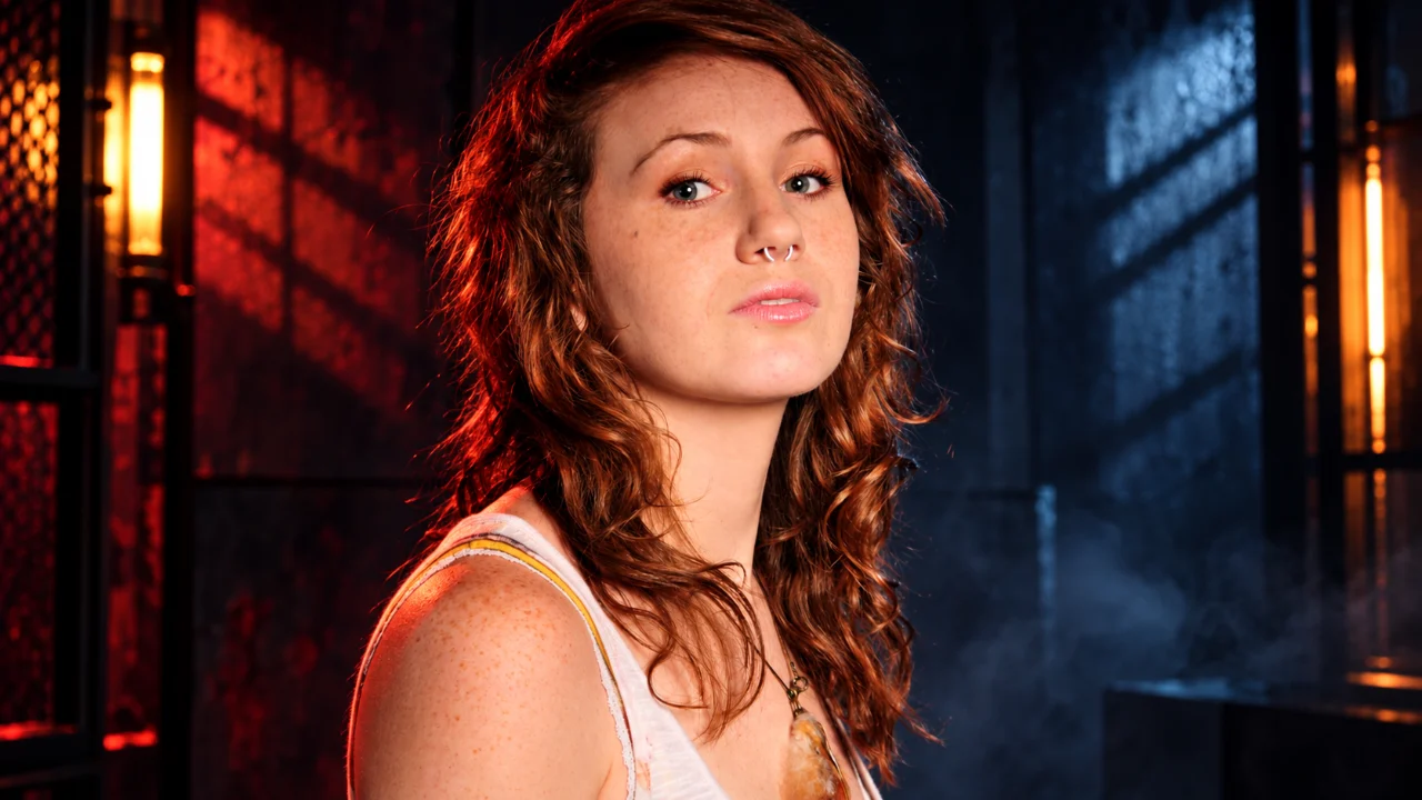 Close-up of a freckled woman with wavy auburn hair, a silver septum ring, and a pale floral tank top. She looks toward the camera in a dark industrial setting illuminated by red and blue lights.