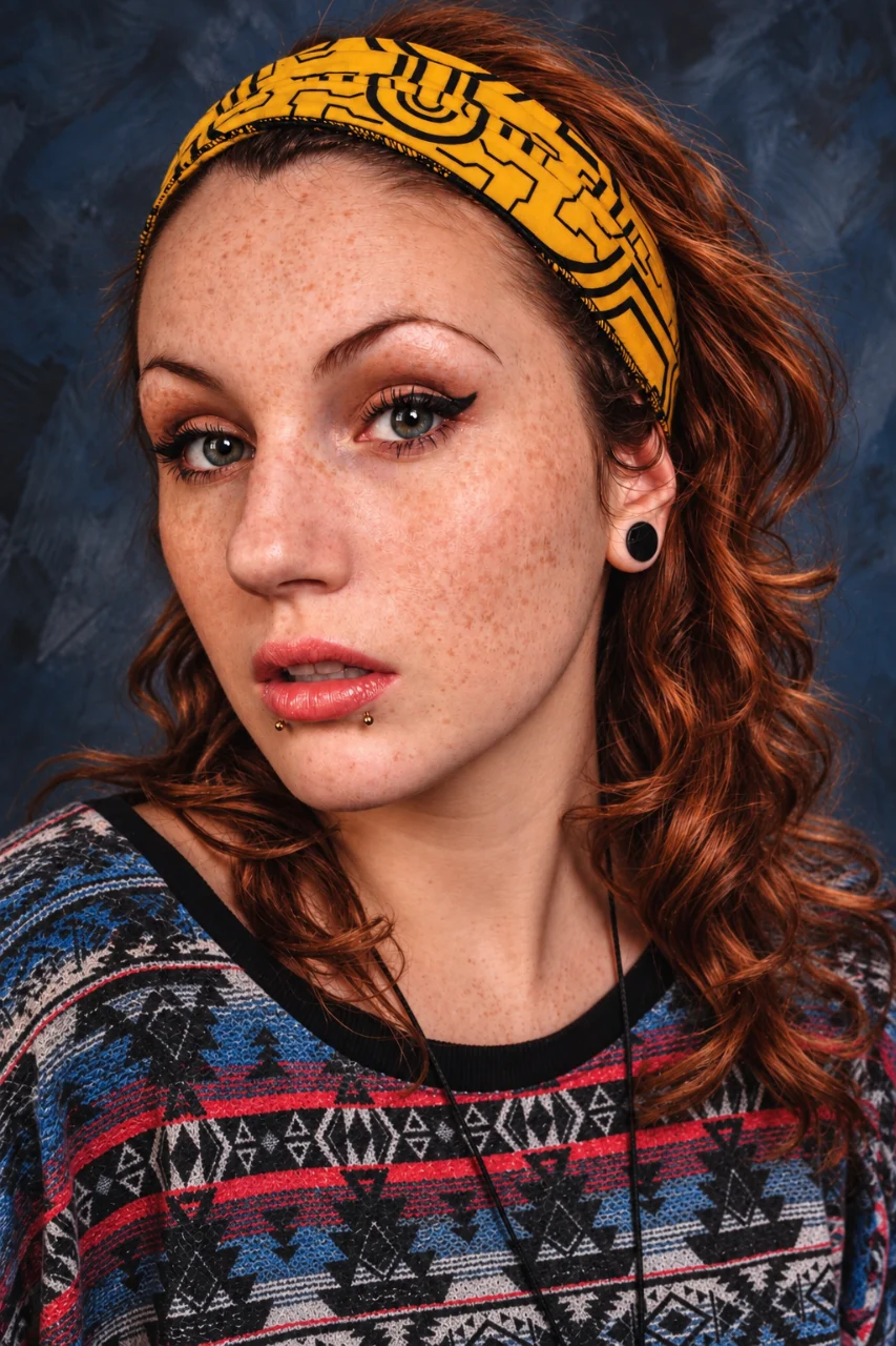 Close-up portrait of a freckled woman with auburn hair, gray-green eyes, black ear gauges, and lower-lip piercings. She wears a yellow patterned headband and a multicolored geometric sweater against a dark blue studio background.