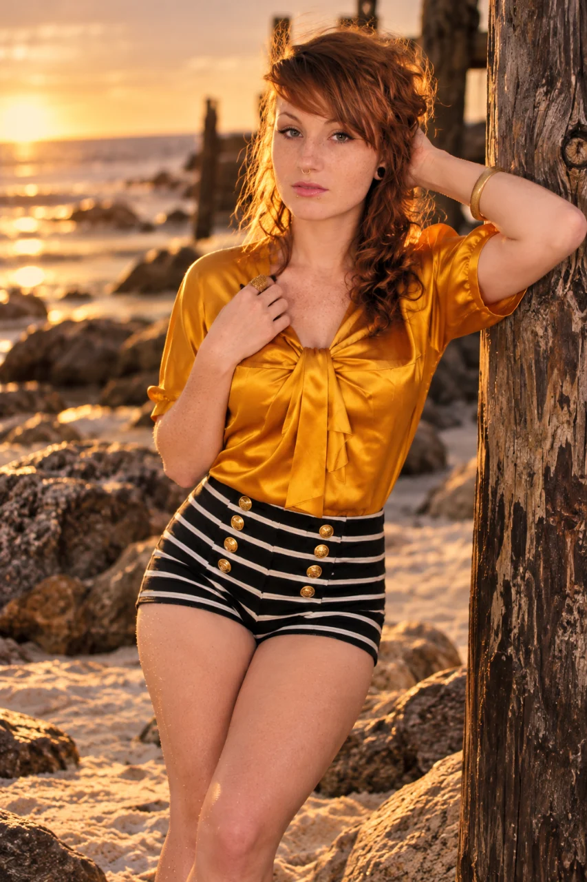 A red-haired woman in a golden blouse and black-and-white striped shorts poses beside a weathered wooden post on a rocky beach at sunset.
