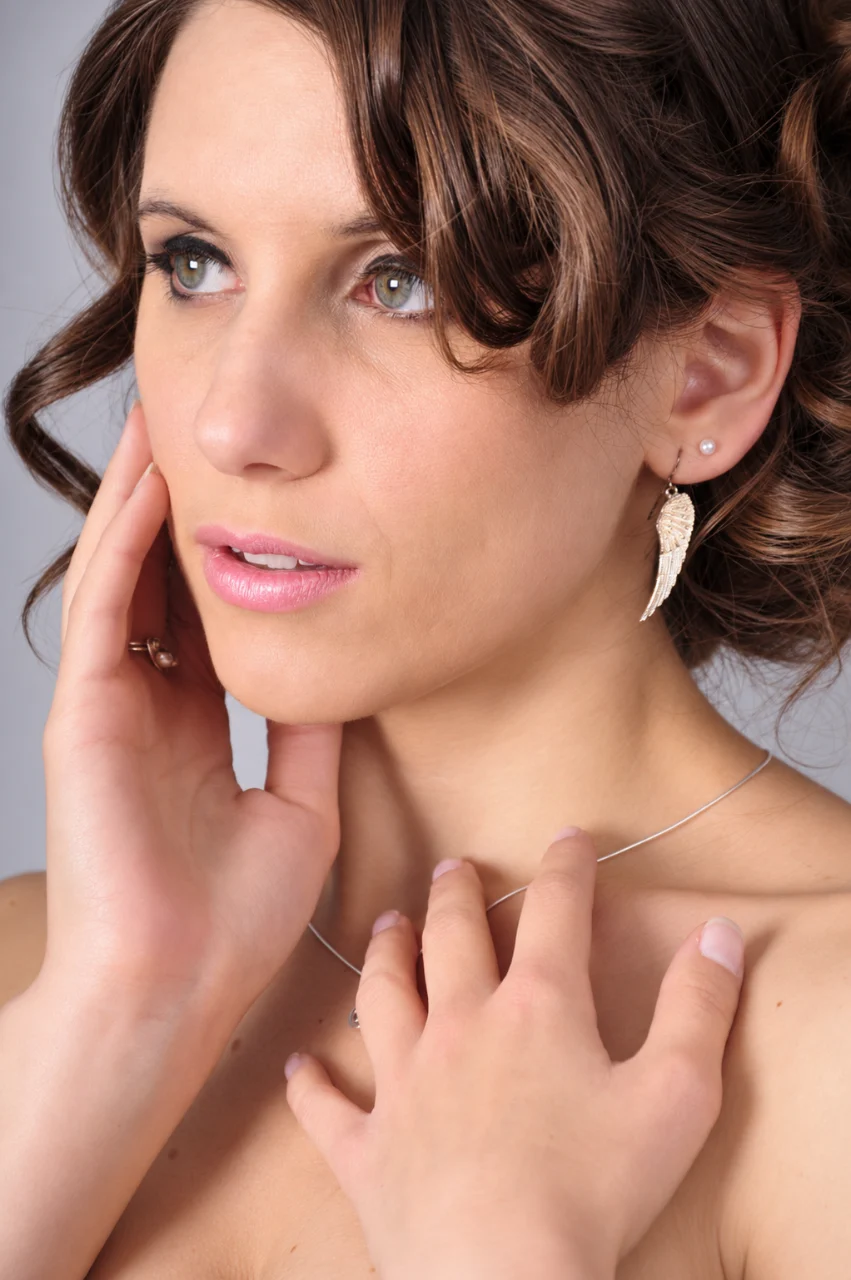 Close-up portrait of a brown-haired woman looking upward, wearing silver feather earrings and a thin necklace, with one hand touching her cheek and the other resting near her collarbone.