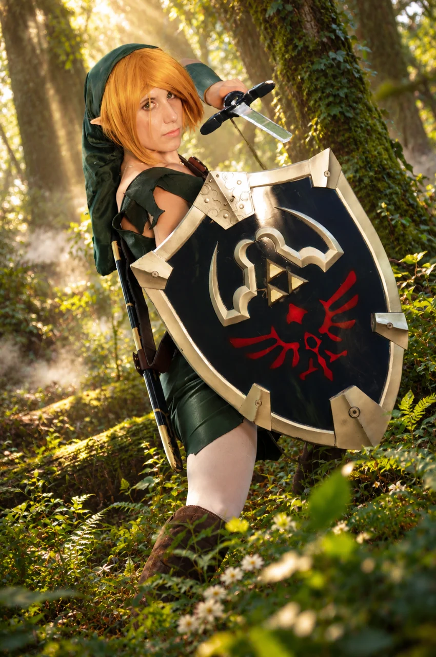Woman dressed as a fantasy elf warrior in green clothing, holding a sword and a large black-and-silver shield with red markings in a sunlit forest.
