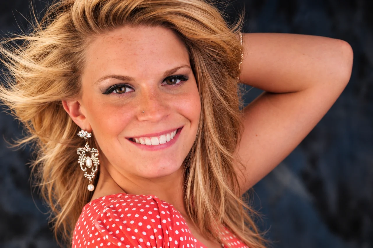 A smiling blonde woman with windswept hair wears a red polka-dot top and ornate earrings against a dark background.