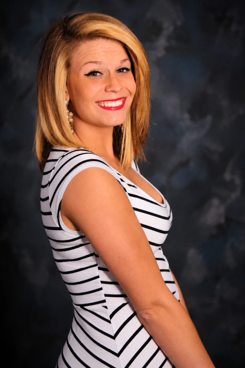 A smiling blonde woman in a black-and-white striped dress poses against a dark blue-gray studio backdrop.