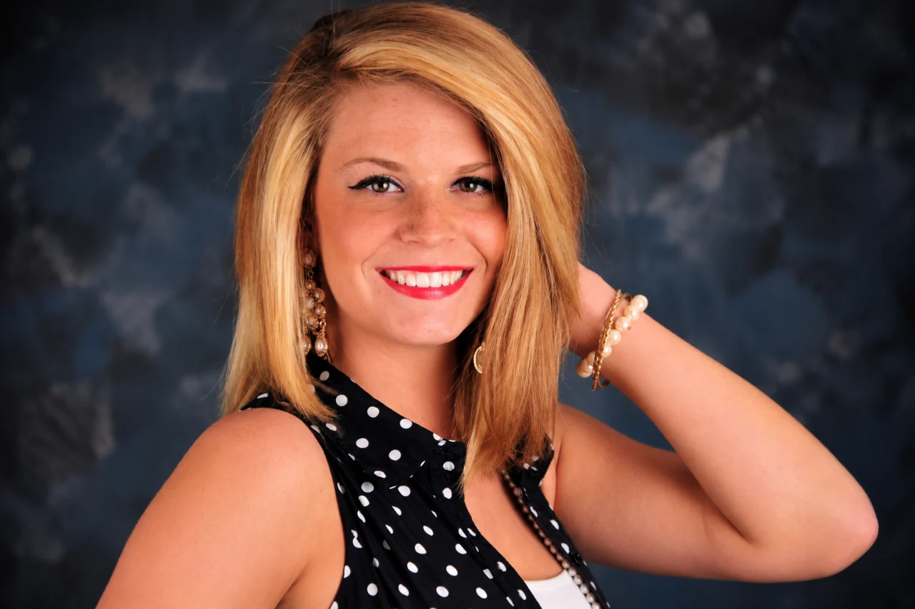 A smiling blonde woman wears a black polka-dot blouse, pearl jewelry, and red nail polish against a dark studio background.