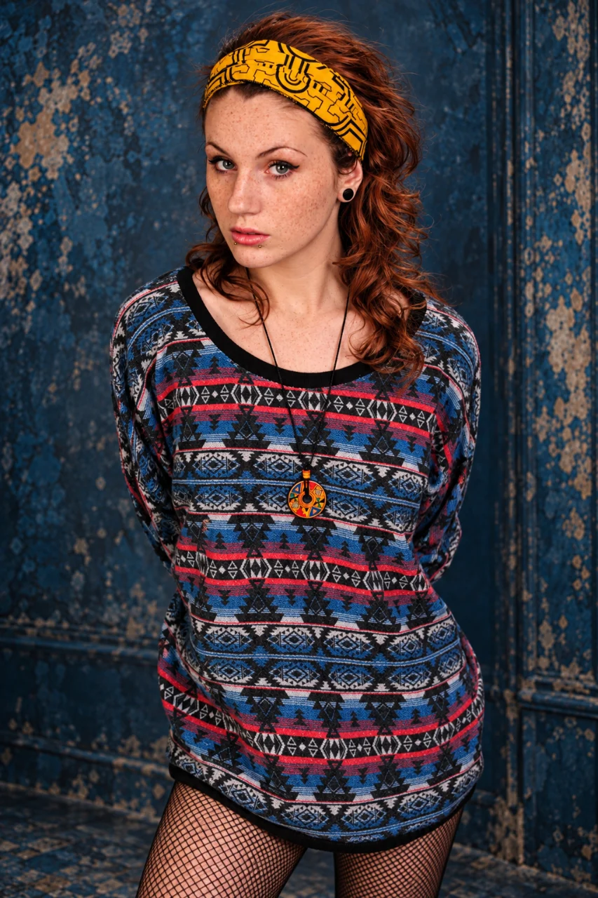 A freckled woman with curly auburn hair stands in front of a peeling blue wall, wearing a yellow patterned headband, oversized blue-and-red geometric sweater, pendant necklace, and fishnet tights. Her hands are held behind her back.