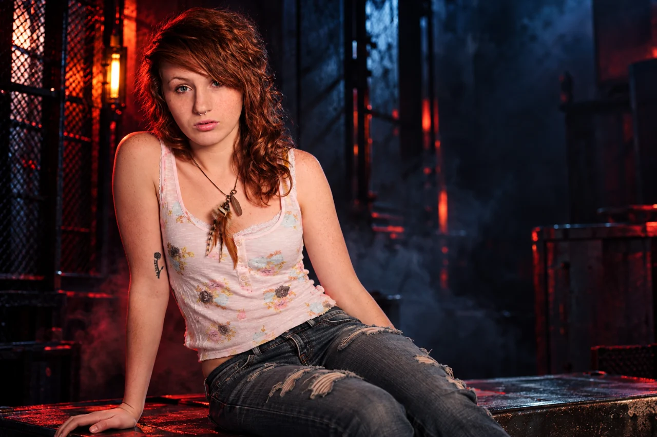 A freckled auburn-haired woman sits on a metal platform in a dark industrial room, wearing a pale floral tank top and ripped blue jeans. Red and blue lighting illuminates the scene, with metal grating and glowing fixtures behind her.