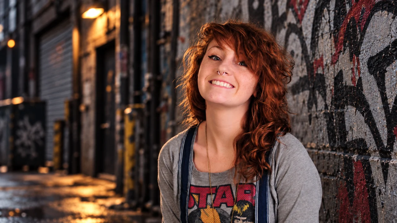 A red-haired woman smiles while leaning against a graffiti-covered wall in an urban alley. She wears a gray Star Wars shirt, striped overalls, and a long necklace, with softly blurred industrial buildings in the background.